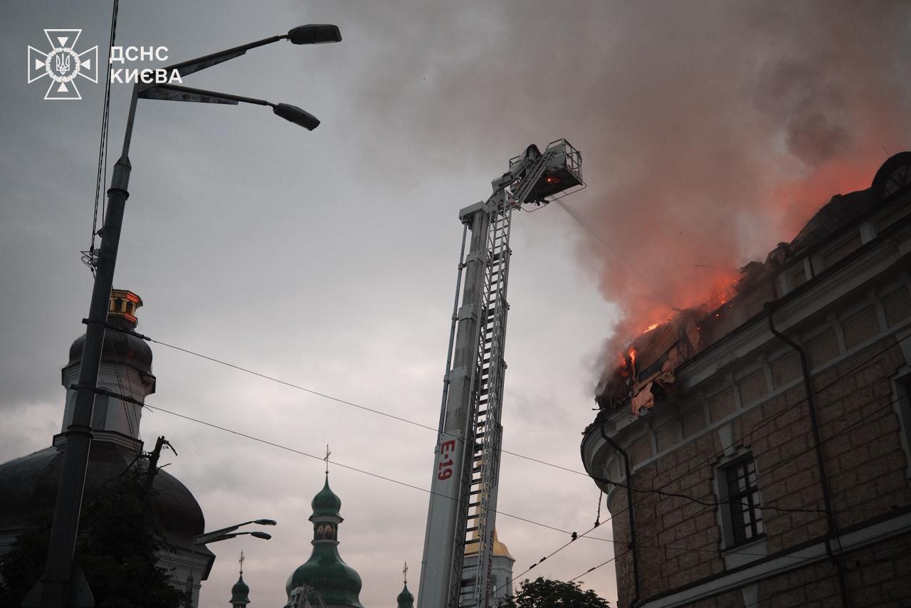 A fire-service aerial ladder sprays the burning corner roof of the Mystetskyi Arsenal at dawn, green Lavra church domes in the foreground, 15 June 2026.